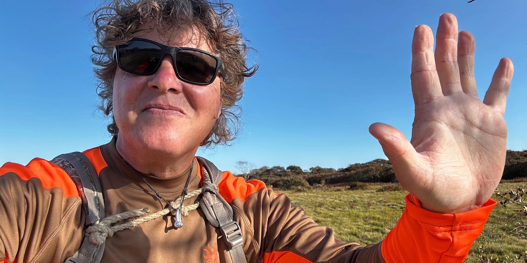 Rich in sunglasses on open moorland, one hand raised to the camera.