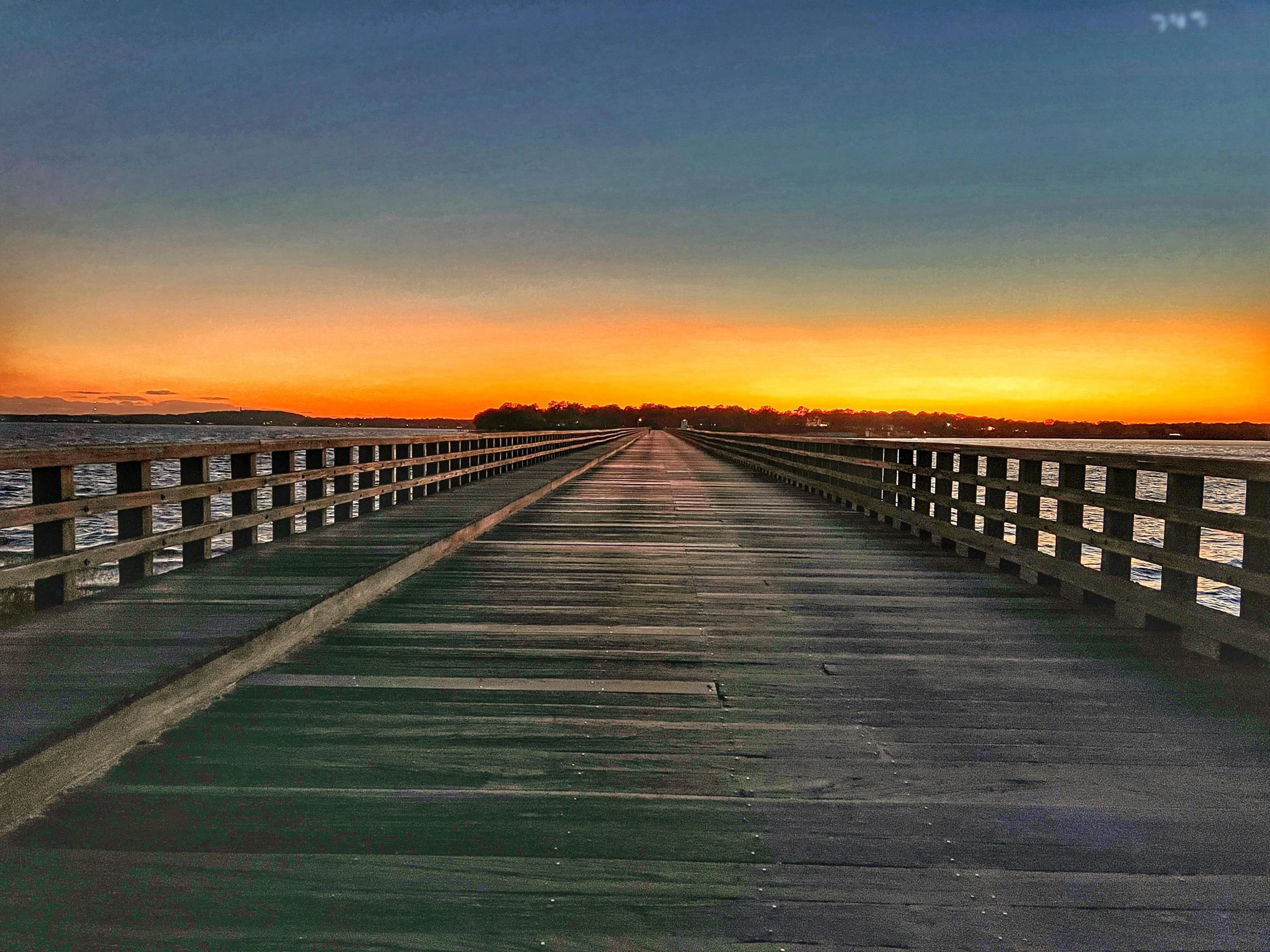 A long wooden bridge running straight out over water toward an orange sunset.