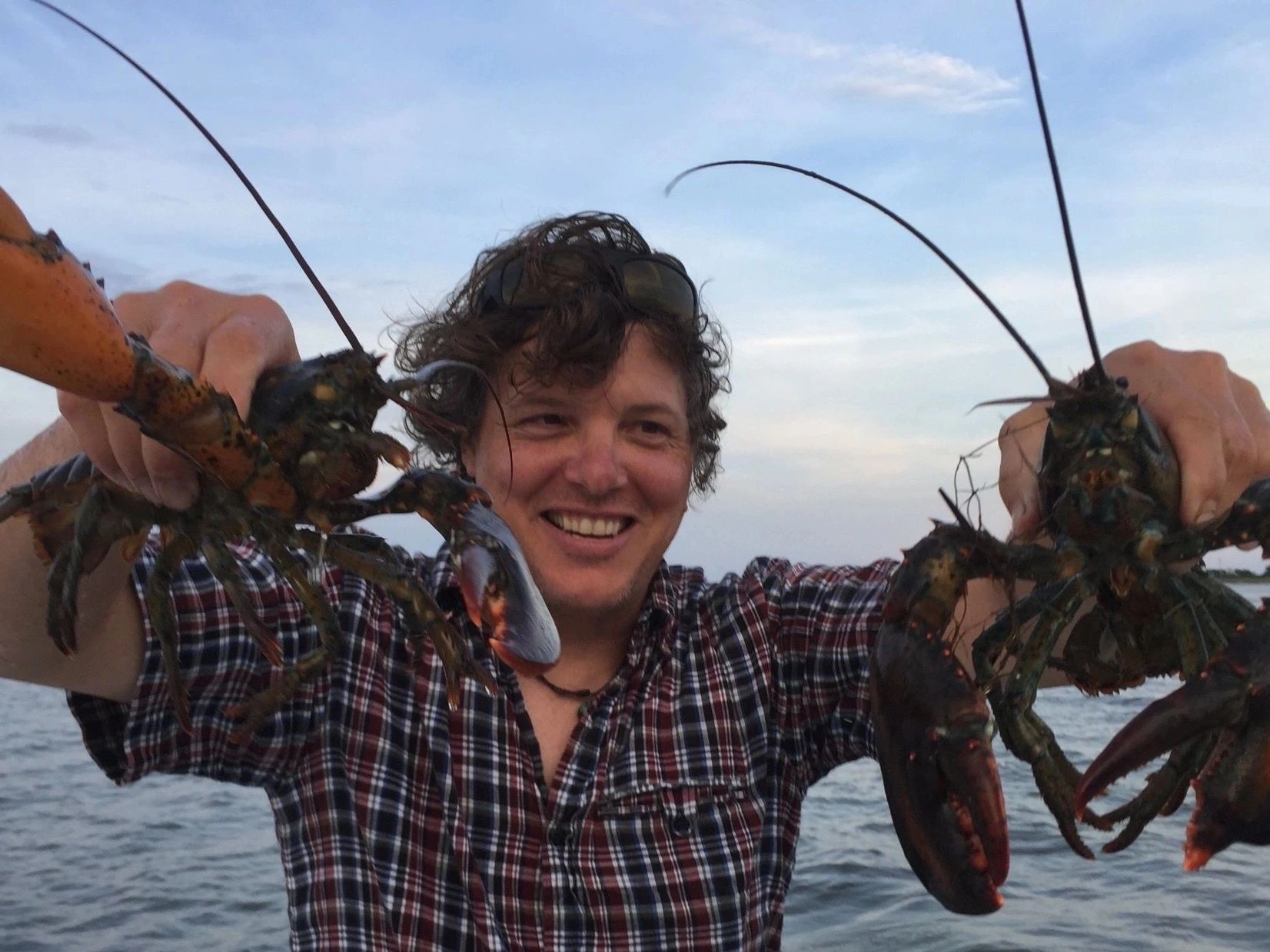 Rich grinning, holding up a lobster in each hand on the water at dusk.