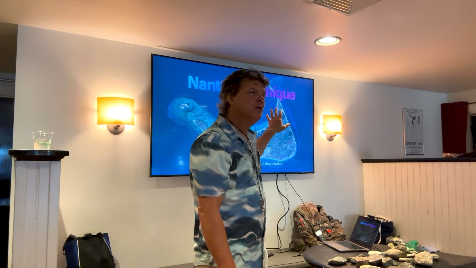 Rich speaking in front of a wall screen showing a Nantucket Unique title slide, rock specimens laid out on a table beside him.