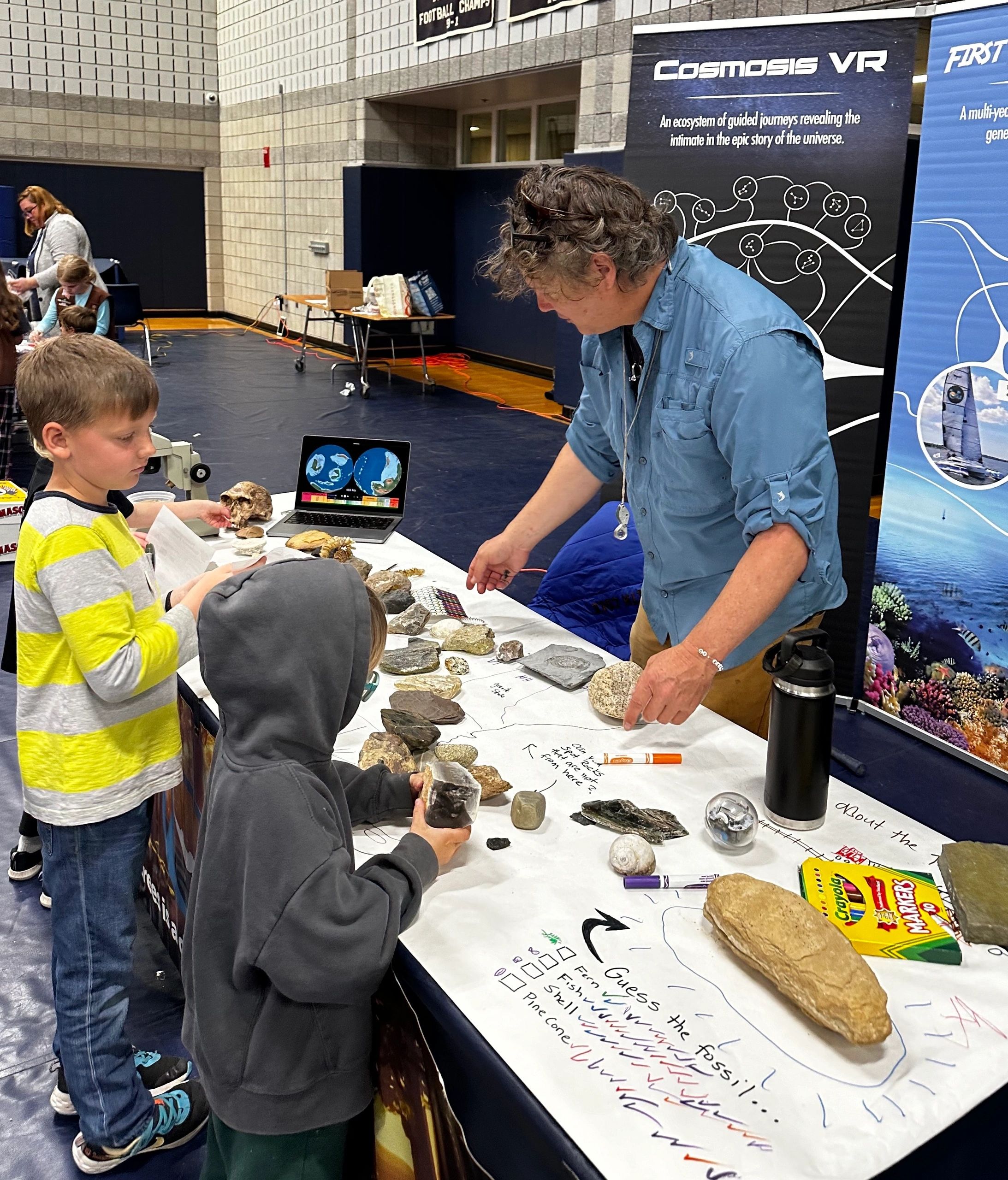 Rich leaning over a table of fossils and rock specimens, talking with two children at a public science event.