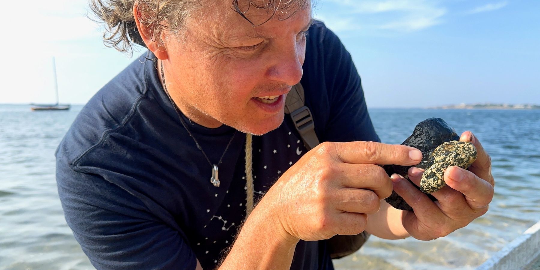 Rich examining two stones in his hands at the water's edge, a moored sailboat behind him.