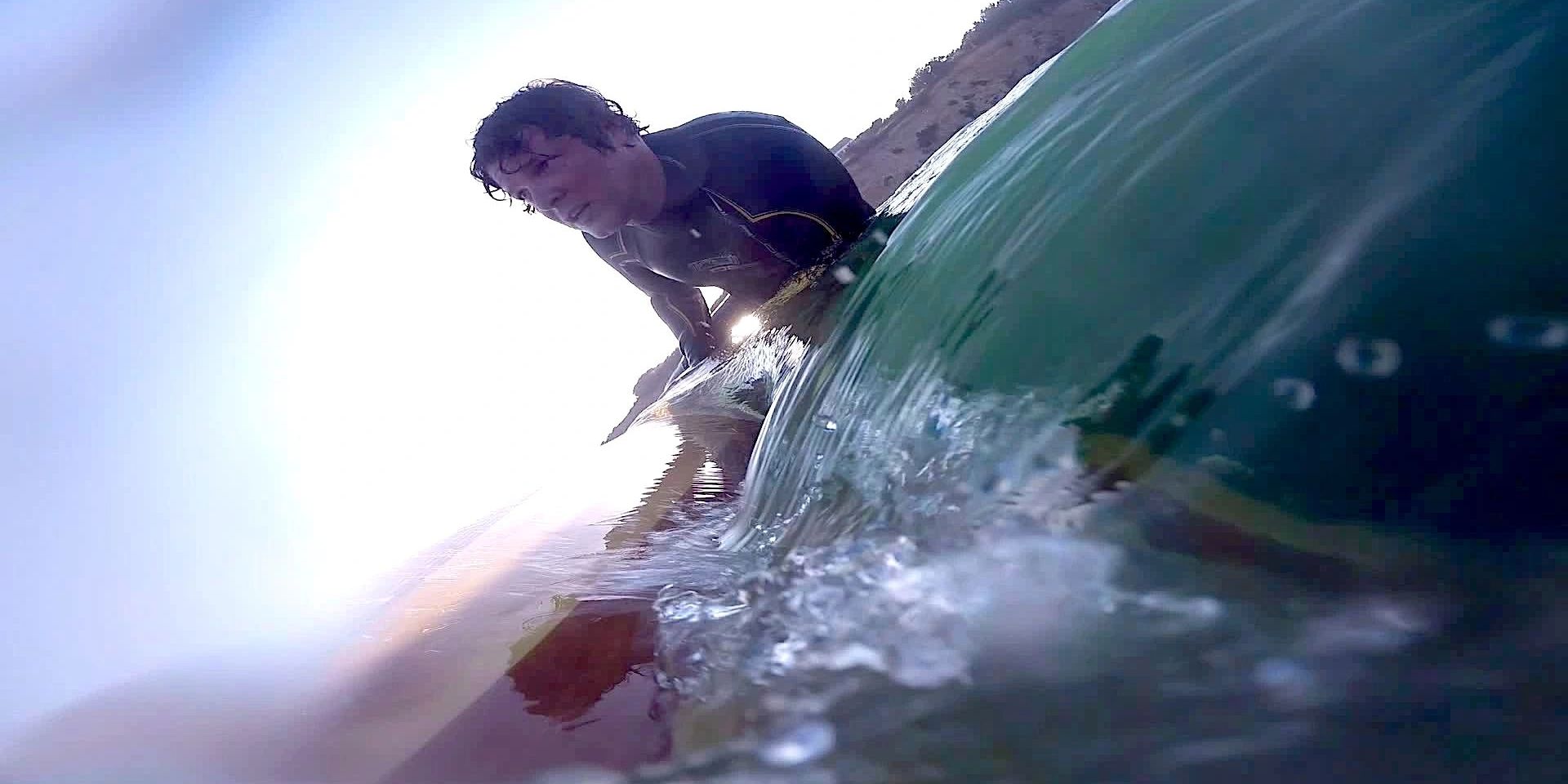 Water-level view of Rich paddling a wooden board as a wave curls across the lens.
