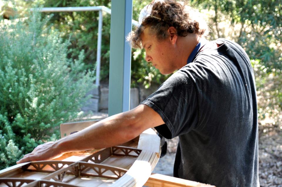 Rich sanding the deck of a wooden surfboard outdoors, the ribbed internal frame visible along the rail.