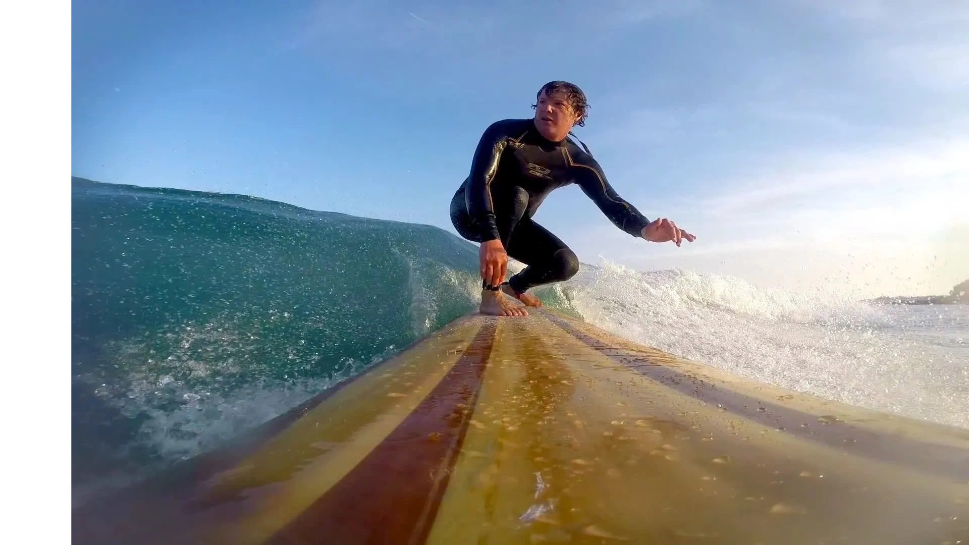 Rich riding a wave crouched on a wooden surfboard, the grain of the deck visible.